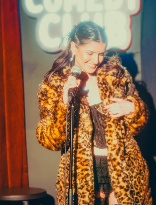 Tori Morancay performing stand-up in a leopard-print coat at a comedy club