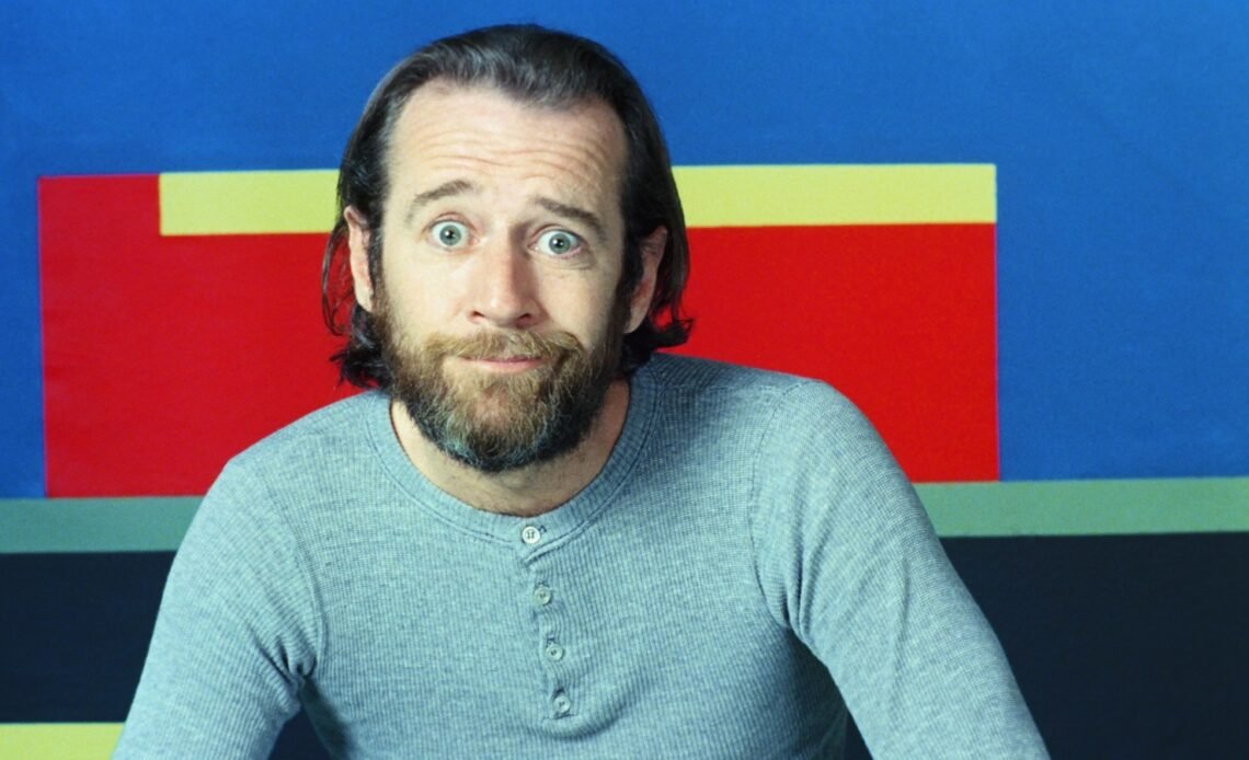 George Carlin with a beard in the 1970s, sitting in front of a blue, red and yellow backdrop